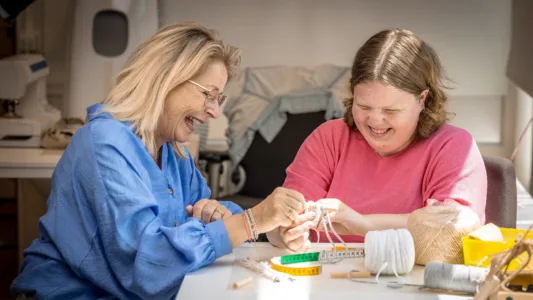 Twee mensen bezig met breien of haken aan een tafel met verschillende benodigdheden zoals garen, meetlint en breinaalden.