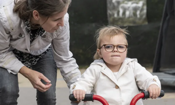 Volwassene leunt over een kind dat op een rode driewieler zit.