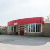 Een bakstenen gebouw met één verdieping en een gebogen rode gevel. Het heeft grote ramen die een rode deur flankeren. Een paar auto's staan geparkeerd aan de linkerkant van het gebouw onder een gedeeltelijk bewolkte lucht.