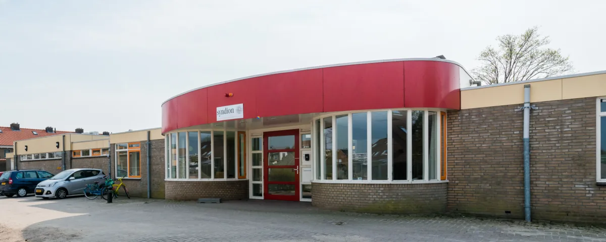 Een bakstenen gebouw met één verdieping en een gebogen rode gevel. Het heeft grote ramen die een rode deur flankeren. Een paar auto's staan geparkeerd aan de linkerkant van het gebouw onder een gedeeltelijk bewolkte lucht.