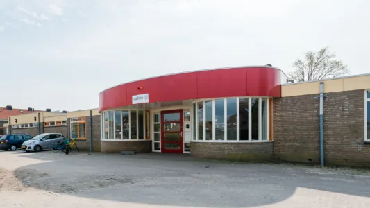 Een bakstenen gebouw met één verdieping en een gebogen rode gevel. Het heeft grote ramen die een rode deur flankeren. Een paar auto's staan geparkeerd aan de linkerkant van het gebouw onder een gedeeltelijk bewolkte lucht.