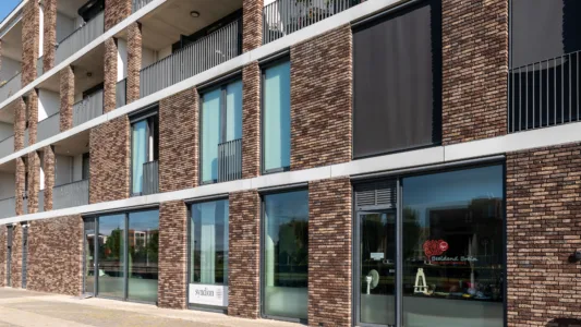 Een modern bakstenen gebouw met grote glazen ramen op de begane grond en balkons erboven. Het gebouw heeft een mix van bruine bakstenen en witte paneelelementen, wat het een eigentijdse uitstraling geeft. De straat ervoor is leeg en zonnig.