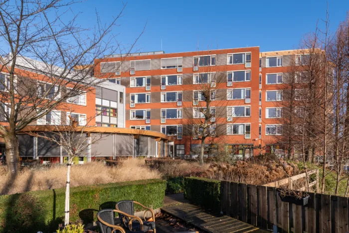 Een rood bakstenen gebouw met meerdere verdiepingen en talloze ramen staat tegen een helderblauwe lucht. Op de voorgrond is een tuin met een houten hek, kale bomen, struiken en een paar stoelen op een terras.
