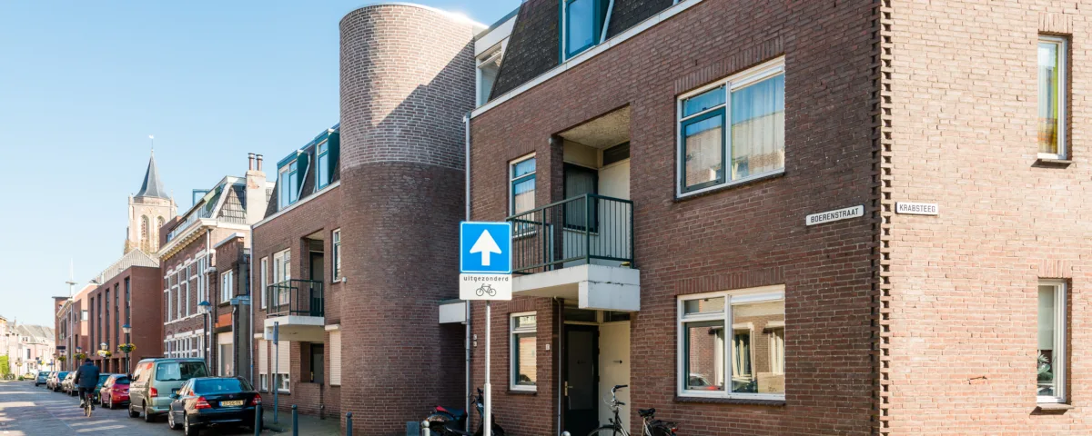 Straatbeeld van een woonwijk met bakstenen gebouwen en een eenrichtingsverkeersbord. Meerdere auto's staan geparkeerd langs de stoep. De zon werpt schaduwen op de grond en de lucht is helder.