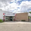 Entree van een gebouw van Syndion met een bord met het logo boven de ingang, bankje en stoelen buiten, naast een lage bakstenen muur en groenvoorziening.