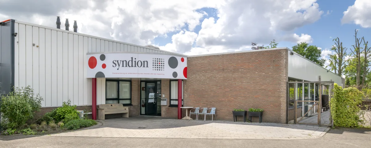 Entree van een gebouw van Syndion met een bord met het logo boven de ingang, bankje en stoelen buiten, naast een lage bakstenen muur en groenvoorziening.