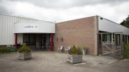 Voorkant van een gebouw met een ingang onder een gebogen luifel waarop 'Syndion' staat. Twee planten staan naast de deur