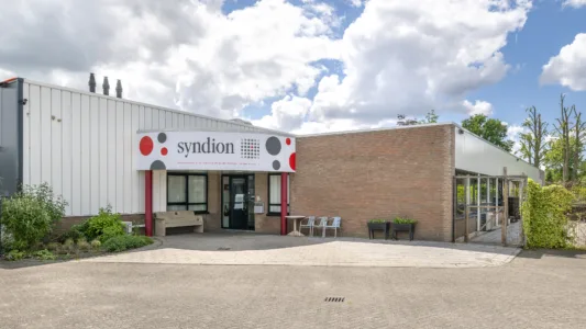 Entree van een gebouw van Syndion met een bord met het logo boven de ingang, bankje en stoelen buiten, naast een lage bakstenen muur en groenvoorziening.