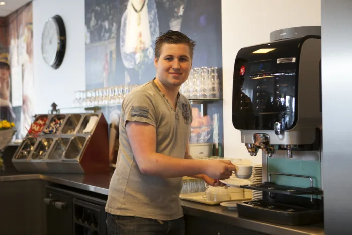 Persoon bedient een koffiemachine, houdt een kopje onder de uitloop. Diverse items op de toonbank, zoals kopjes en apparatuur.