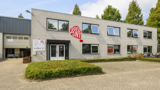 Twee verdiepingen tellend gebouw met witte bakstenen gevel, ramen, en een bord met "GoedPunt" in rode letters. Gebied voor het gebouw bestraat met stenen, groene struiken langs de rand. Bomen op de achtergrond.