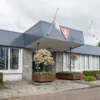 Vooraanzicht van een modern bedrijfsgebouw met een overkapping bij de ingang, vlaggen aan de gevel en plantenbakken met kleine bomen langs het trottoir.