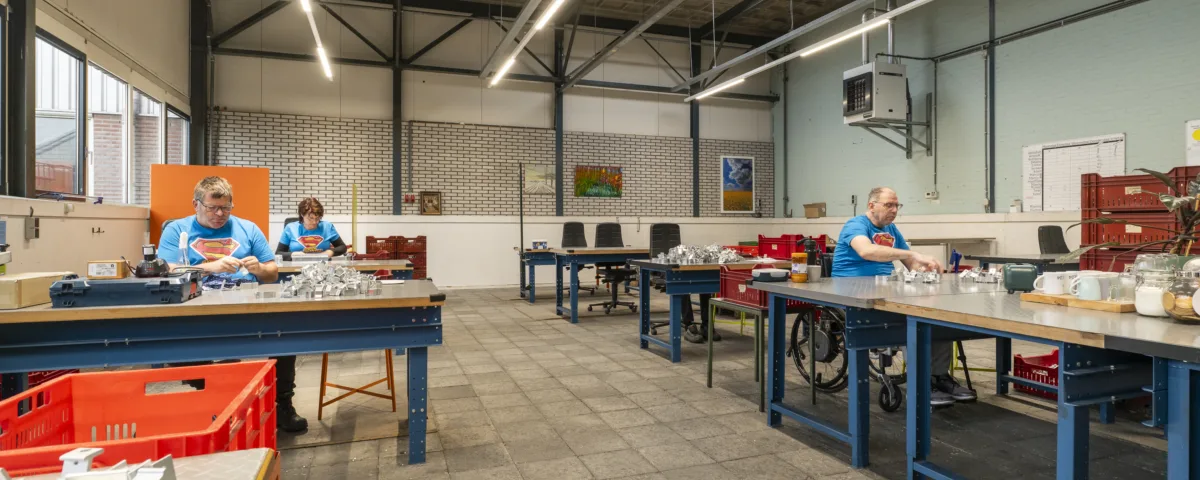 Werkruimte waar meerdere mensen aan tafels kleine onderdelen sorteren en monteren, met bakken, gereedschap en werkbanken in een grote hal.