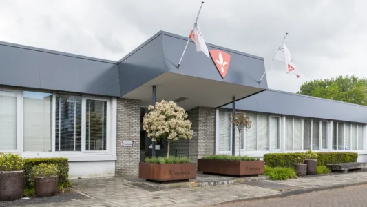 Vooraanzicht van een modern bedrijfsgebouw met een overkapping bij de ingang, vlaggen aan de gevel en plantenbakken met kleine bomen langs het trottoir.
