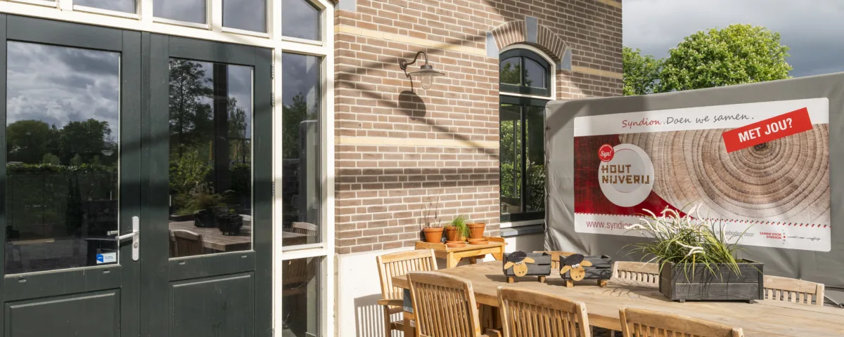 Buitenplaats bij een bakstenen gebouw met een deur en ramen, ingericht met een houten tafel en stoelen, planten en een bord met informatie over houtnijverij.
