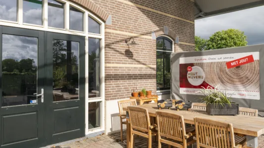 Buitenplaats bij een bakstenen gebouw met een deur en ramen, ingericht met een houten tafel en stoelen, planten en een bord met informatie over houtnijverij.