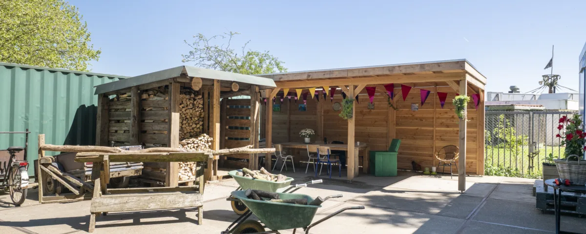Buitenruimte met houten overkapping en houtopslag, ingericht met tafel en stoelen, kruiwagens en stapels hout op een zonnige dag.