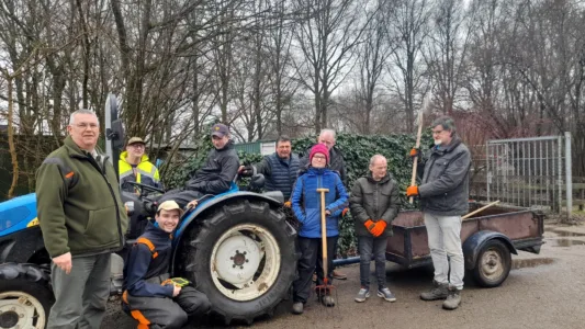 Een groep mensen in werkkleding voor buiten staat rond een blauwe tractor met aanhanger. Ze houden tuingereedschap vast en lachen naar de camera. Bomen zonder bladeren zijn zichtbaar op de achtergrond.