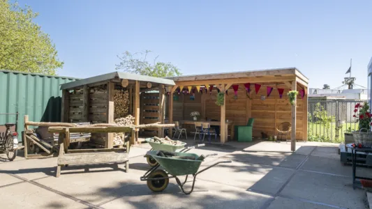 Buitenruimte met houten overkapping en houtopslag, ingericht met tafel en stoelen, kruiwagens en stapels hout op een zonnige dag.