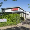 Gebouw van het NAH Centrum met een entree langs een pad, omgeven door groen en voorzien van een duidelijk zichtbaar naam- en logobord aan de gevel.