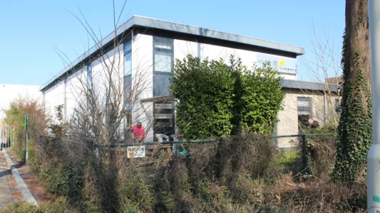 Een klein, modern gebouw met een plat dak, omgeven door groen en een hek. Er is een bosrijk gebied aan de voorkant en een helderblauwe lucht erboven.