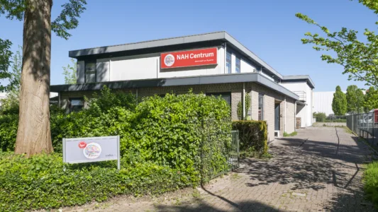 Gebouw van het NAH Centrum met een entree langs een pad, omgeven door groen en voorzien van een duidelijk zichtbaar naam- en logobord aan de gevel.