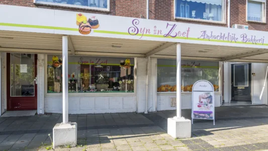 Gevel van een winkelpand met de naam “Syn in Zoet Ambachtelijke Bakkerij”, met etalages vol bakproducten en een stoepbord bij de ingang onder een overkapping.