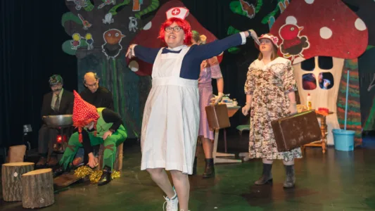 Een groep mensen op een podium, gekleed in kostuums. De achtergrond bevat grote, kleurrijke rekwisieten die op bomen en paddenstoelen lijken. Eén persoon op de voorgrond draagt een wit schort en een hoed met een rood kruis en strekt zijn armen uit. Andere personen zijn gekleed in verschillende outfits, waaronder bloemenjurken en hoeden. Er zitten ook twee mensen aan de linkerkant van het podium, waarvan één een groen kostuum met een rode hoed lijkt te dragen.
