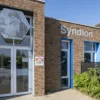 Entree van een bakstenen gebouw van Syndion met glazen deur, blauwe kozijnen en een kunstwerk boven de ingang, met naast de deur een bord van kinderdagcentrum Schuttersplein.