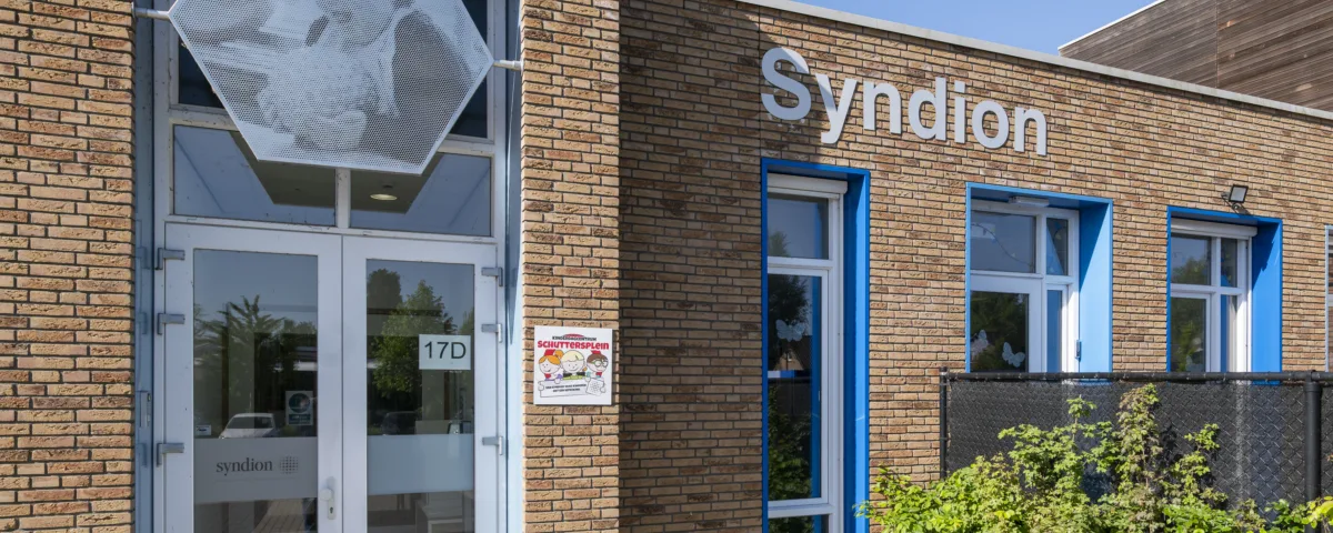 Entree van een bakstenen gebouw van Syndion met glazen deur, blauwe kozijnen en een kunstwerk boven de ingang, met naast de deur een bord van kinderdagcentrum Schuttersplein.