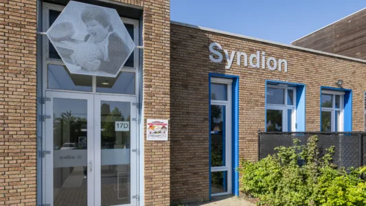 Entree van een bakstenen gebouw van Syndion met glazen deur, blauwe kozijnen en een kunstwerk boven de ingang, met naast de deur een bord van kinderdagcentrum Schuttersplein.