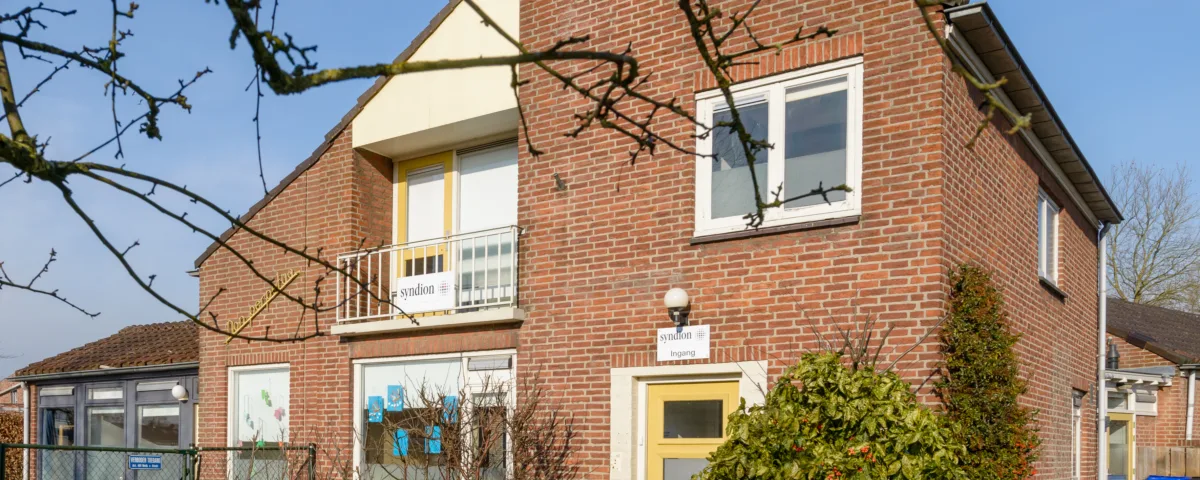 Twee verdiepingen tellend bakstenen huis met gele voordeur en witte ramen, balkon met gele reling, en struiken ervoor.