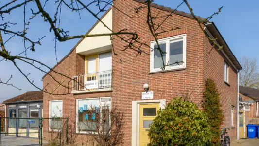 Twee verdiepingen tellend bakstenen huis met gele voordeur en witte ramen, balkon met gele reling, en struiken ervoor.
