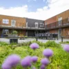 Binnenplaats van een woongebouw met bakstenen gevels en galerijen, ingericht met plantenbakken, zitplekken en een lange tafel met stoelen.