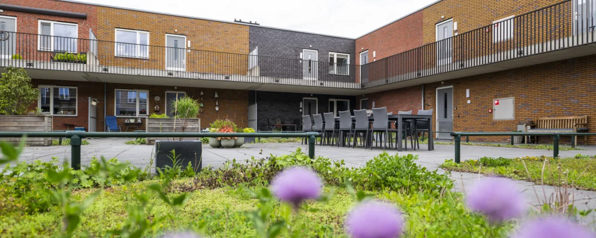 Binnenplaats van een woongebouw met bakstenen gevels en galerijen, ingericht met plantenbakken, zitplekken en een lange tafel met stoelen.