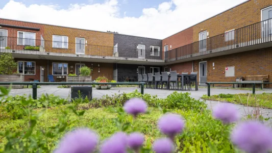 Binnenplaats van een woongebouw met bakstenen gevels en galerijen, ingericht met plantenbakken, zitplekken en een lange tafel met stoelen.