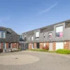 Moderne woonlocatie met twee aaneengeschakelde gebouwen van baksteen en donkere gevelpanelen rond een bestraat binnenplein, met zitbank en groen, onder een heldere blauwe lucht.