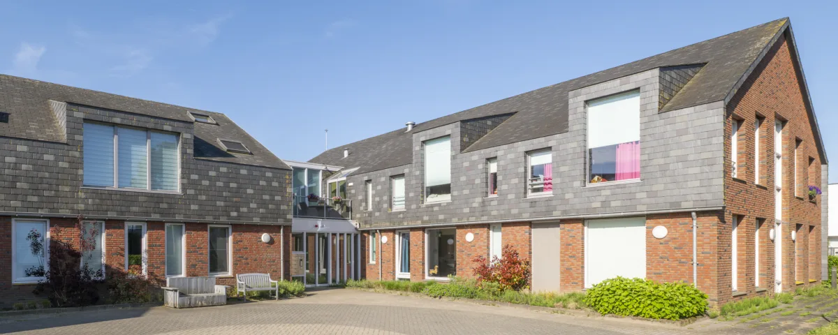 Moderne woonlocatie met twee aaneengeschakelde gebouwen van baksteen en donkere gevelpanelen rond een bestraat binnenplein, met zitbank en groen, onder een heldere blauwe lucht.