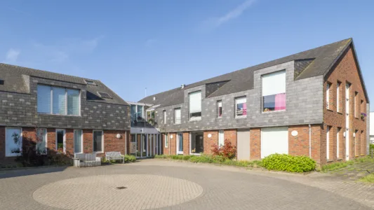 Moderne woonlocatie met twee aaneengeschakelde gebouwen van baksteen en donkere gevelpanelen rond een bestraat binnenplein, met zitbank en groen, onder een heldere blauwe lucht.