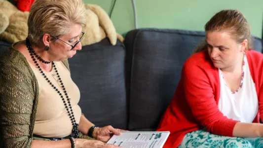 Twee mensen zitten op een bank. Eén persoon houdt een open boek of tijdschrift vast en beiden lijken in gesprek te zijn