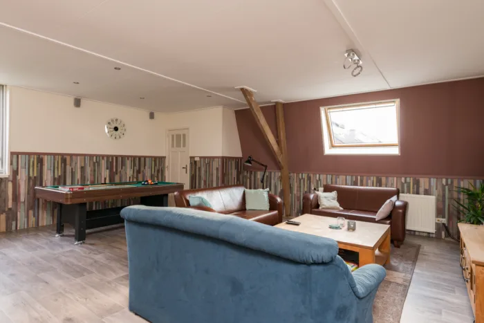 Een gezellige woonkamer met blauwe en bruine leren banken, een houten salontafel, kleurrijke houten panelen op de muren, natuurlijk licht van een raam en een pooltafel
