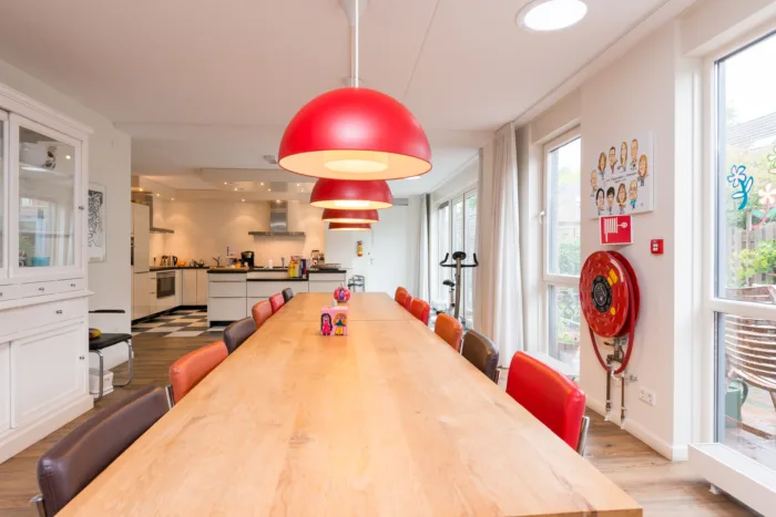 Moderne eetruimte met lange houten tafel, kleurrijke stoelen, rode hanglampen en open keuken
